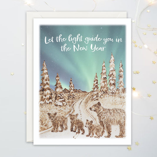 Greeting card illustration with four bears  on a winding path, snowy landscape, Northern Lights and text 'Let the light guide you in the New Year'