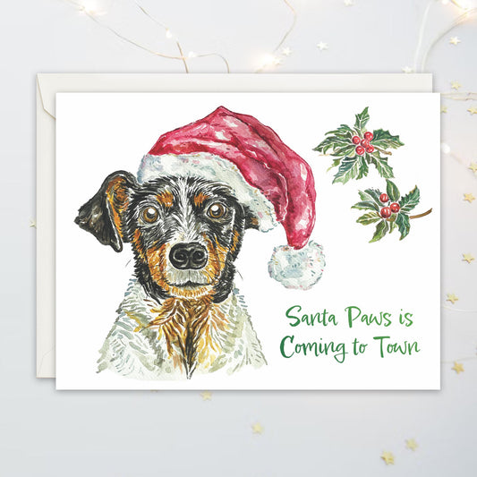 Christmas card with a dog wearing a Santa hat, holly leaves and festive texts "Santa Paws is Coming to Town"