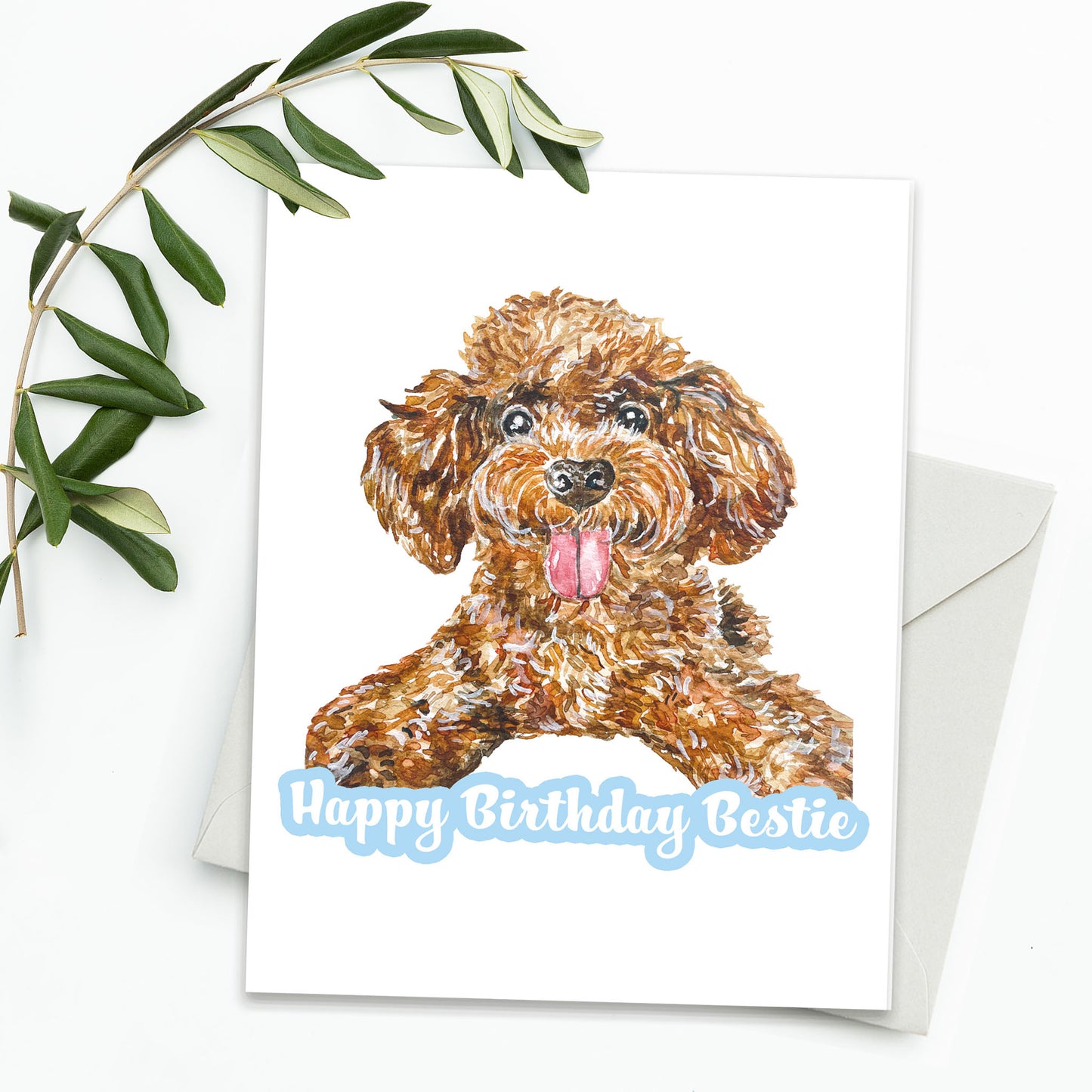 A picture of a greeting card, envelope and green leaves. The card features a brown puppy looking very excited to see you. The text reads: Happy Birthday Bestie!