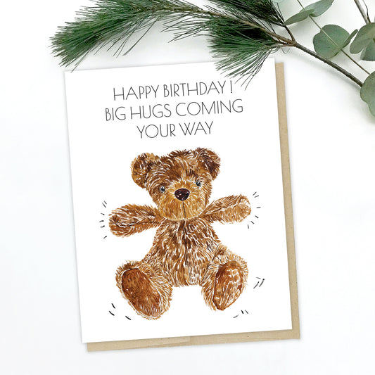 A greeting card with pine leaves in the background. The card has a cute teddy bear waving its arm, looking like it's about to give you a hug. The text reads: Happy Birthday! Big Hugs Coming Your Way.