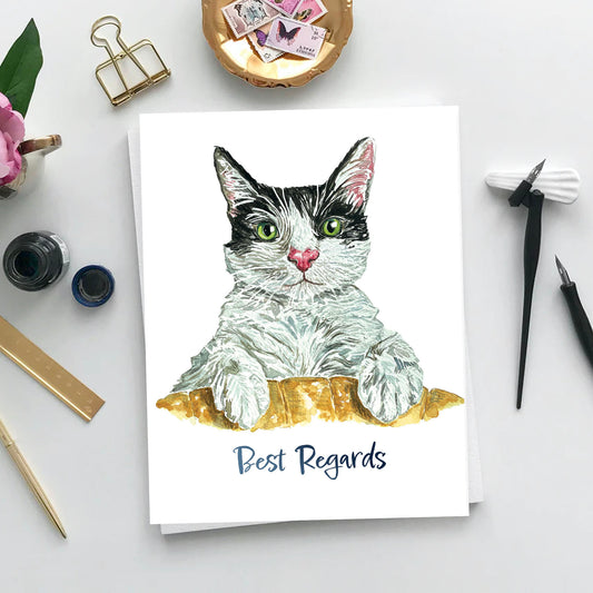 A greeting card on a table with some stamps, pen, and ink. The card has a black and white cat with green eyes looking at you intensely. It is resting its paws on a chair. The text simply reads: Best Regards.