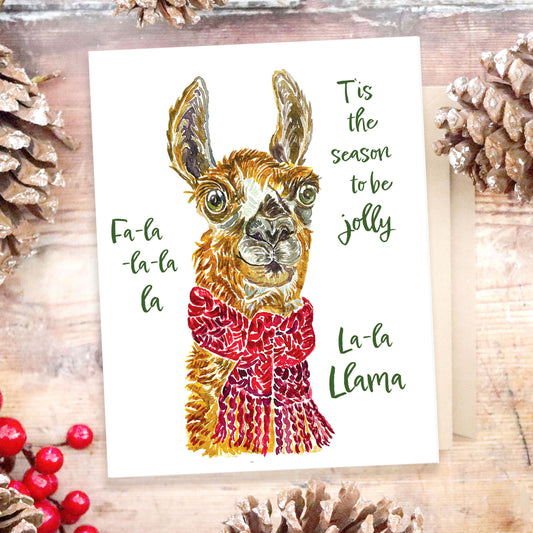 An image of a holiday greeting card with pinecones in the background. The card features a brown llama with green eyes, his ears are tall like bananas, and he is wearing a red scarf around the neck. The text reads: T'is the season to be jolly, Fa la la la la, la la llama.