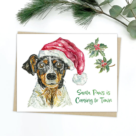 A holiday card of a happy Swiss mountain dog with bright brown eyes, wearing a fluffy santa hat. There are holly and berry branches in the back. The text reads: Santa Paws is Coming to Town.