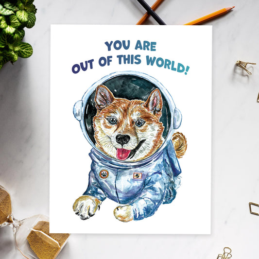 Greeting card of a laughing Shiba Inu puppy wearing a full space suit and helmet. Blue text hovering on top says, You are out of this world!