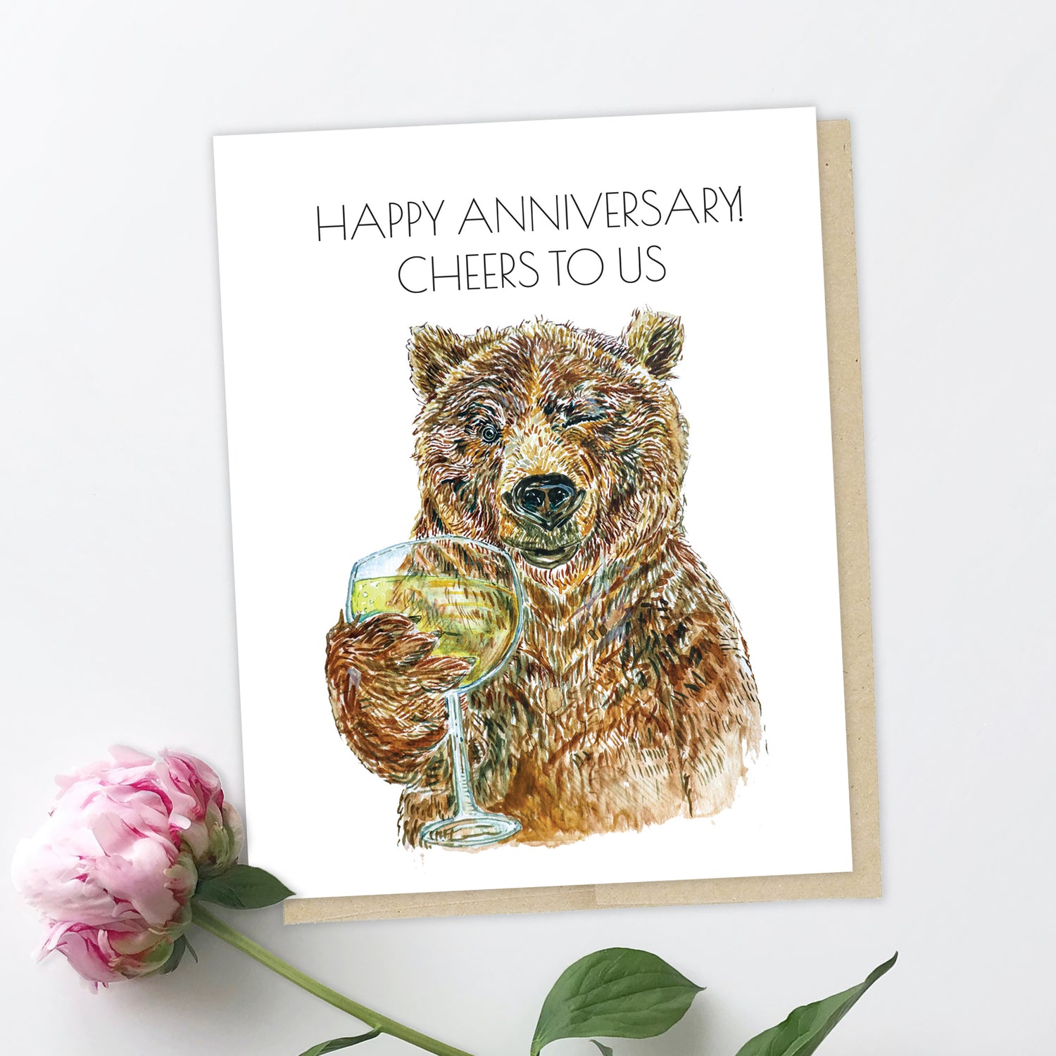 A greeting card and a peony. The card features a brown, grizzly bear holding a glass of champagne. It is winking and looking at you, smiling, the expression like Jay Gatsby in the party scene in the Great Gatsby movie. The text reads: Happy Anniversary! Cheers to us.