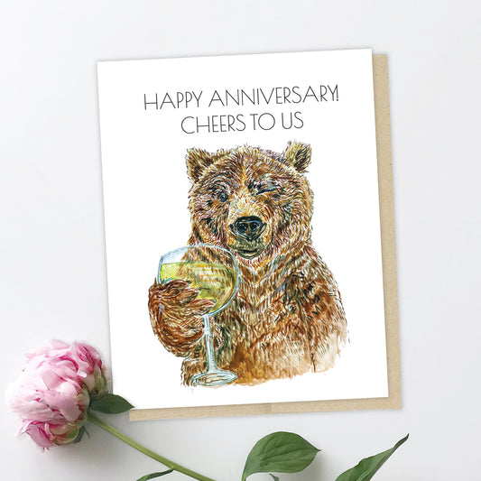 A greeting card and a peony. The card features a brown, grizzly bear holding a glass of champagne. It is winking and looking at you, smiling, the expression like Jay Gatsby in the party scene in the Great Gatsby movie. The text reads: Happy Anniversary! Cheers to us.