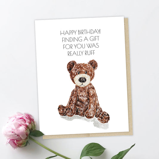 A picture of a greeting card with a little character that looks like a bear dog. He's part bear with his ears, part dog with his snout and paws. He looks cute but a little stressed. The text reads: Happy birthday! Finding you a gift was really "Ruff".