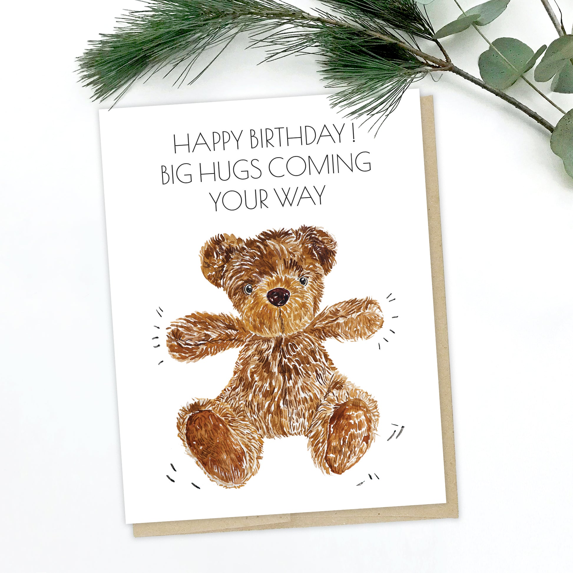 A greeting card with pine leaves in the background. The card has a cute teddy bear waving its arm, looking like it's about to give you a hug. The text reads: Happy Birthday! Big Hugs Coming Your Way.