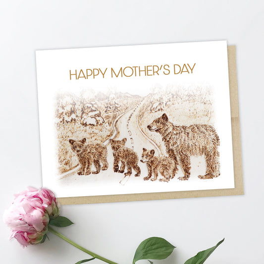 An image of a greeting card with a pink peony in the background. The card features drawing of a family of bears, three little brown grizzly bear cubs and a mama bear, they are crossing the street with trees and mountains in the background. Card is in sepia tone, as it was drawn by pyrography. The text reads: Happy Mother's Day.